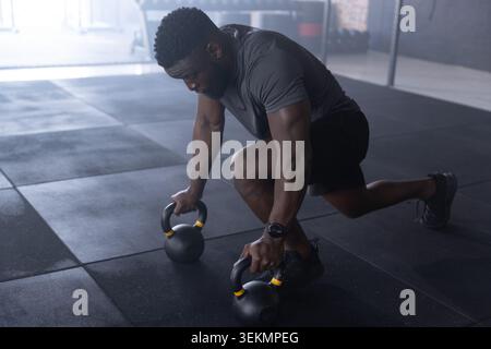 Afroamerikanischer Mann, der auf dem Fußboden des Fitnessstudios eine Kettlebell-Longe vorführt und Kettlebells mit Smartwatch packt Stockfoto
