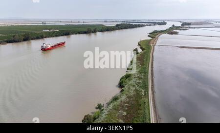 Rotes Frachtboot, das entlang des schlammigen Flusses Guadalquivir in La Puebla del Rio fährt, vorbei an grünen landwirtschaftlichen Feldern und überfluteten Reisfeldern Stockfoto