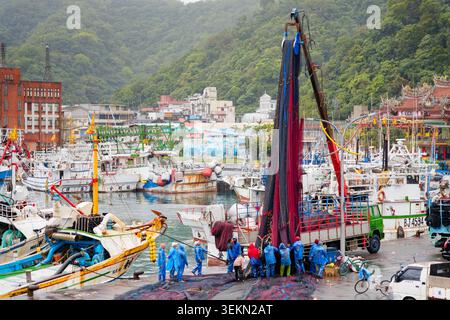 Yilan, Taiwan - 31. März 2025: Besatzung in Regenmänteln hilft, ein Schleppnetz mit Kran auf ein Fischereischiff in Nanfangao zu laden, hinter dem die Hafenstadt liegt. Stockfoto