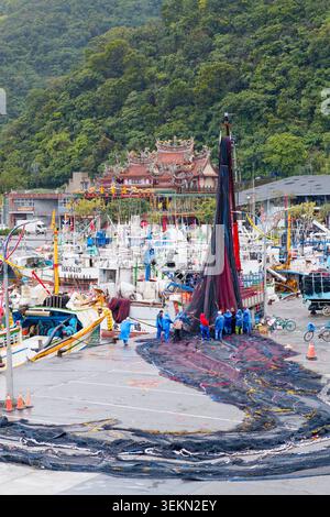 Yilan, Taiwan - 31. März 2025: Die Besatzung von Migranten unterstützt einen Kran beim Heben eines Schleppnetzes im Hafen von Nanfangao mit einem taiwanesischen Tempel und grünen Hügeln dahinter. Stockfoto