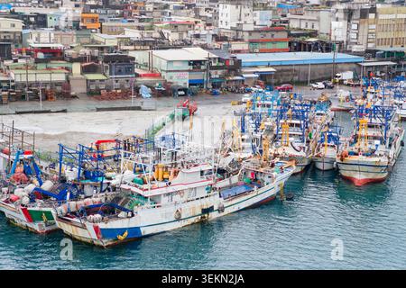 Yilan, Taiwan - 31. März 2025: Fischereifahrzeuge mit blauer und gelber Takelage vertäuten an einem regnerischen Tag im Hafen von Nanfangao mit dichten Stadtgebäuden. Stockfoto
