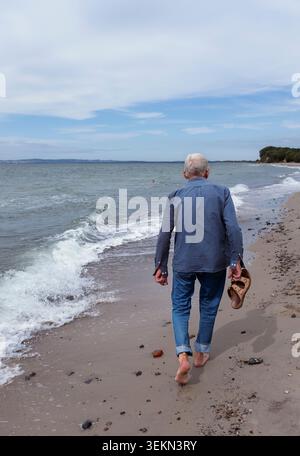 Rückansicht eines älteren Mannes, der Sandalen in der Hand hält und am Strand vor klarem Himmel läuft Stockfoto
