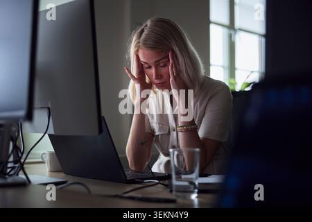 Erschöpfte junge Geschäftsfrau berührt die Tempel, während sie am Schreibtisch im Büro sitzt Stockfoto
