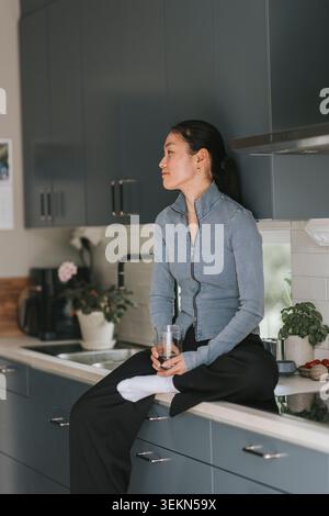 Nachdenkliche junge Frau, die zu Hause mit Trinkglas auf der Küchenzeile sitzt Stockfoto