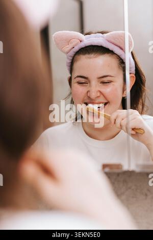 Spiegelreflexion einer lächelnden jungen Frau mit Katzenohren-Stirnband beim Zähneputzen im Badezimmer Stockfoto
