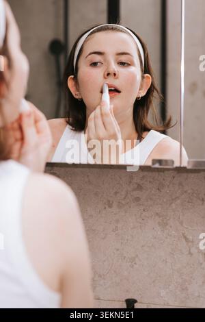 Eine junge Frau, die Lippenbalsam aufträgt, während sie im Spiegel ins Badezimmer blickt Stockfoto