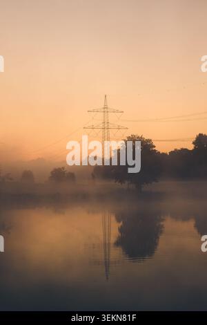 Über einem ruhigen, ländlichen See steht bei Sonnenaufgang ein hoher Strompylon mit weichem Nebel, Silhouettenbäumen und warmem goldenem Licht, das auf dem Wasser reflektiert wird Stockfoto