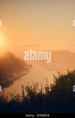Sanftes Morgenlicht erfüllt ein nebeliges Flusstal mit ruhigem Wasser, silbrigem Schilf, fernen Bäumen und sanften Hügeln unter einem warmen orangen Himmel Stockfoto
