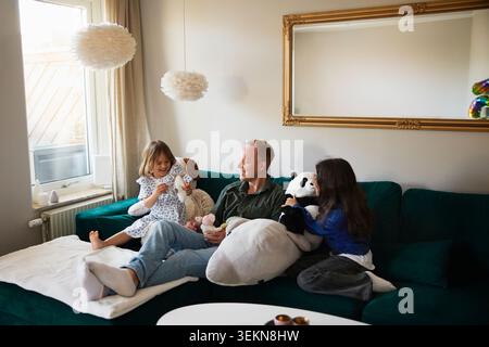 Hochwinkelansicht des Vaters, der mit Töchtern sitzt, die zu Hause mit Spielzeug auf dem Sofa spielen Stockfoto