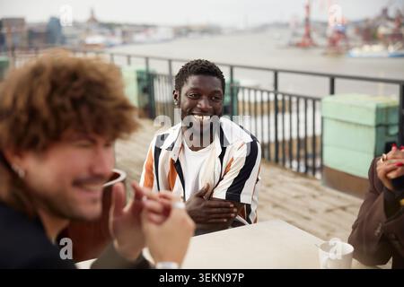 Lächelnder junger Unternehmer, der während der Kaffeepause mit Kollegen auf der Büroterrasse sitzt Stockfoto
