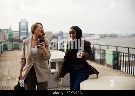 Glückliche, multirassische Unternehmerinnen, die Kaffeetassen halten, während sie auf der Büroterrasse laufen Stockfoto