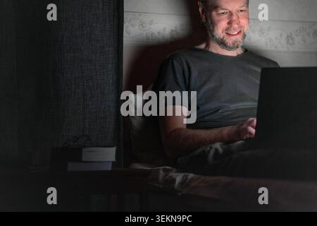 Lächelnder Mann mit Laptop, während er zu Hause auf dem Bett saß Stockfoto