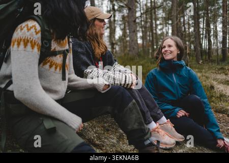 Lächelnde Frau, die Freizeit mit Freundinnen verbringt, während sie auf dem Boden im Wald sitzt Stockfoto