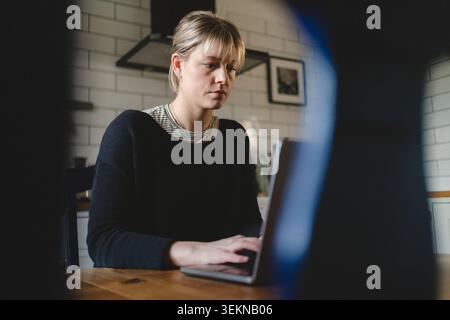 Konzentrierte blonde Geschäftsfrau, die am Laptop arbeitet, während sie zu Hause am Tisch sitzt Stockfoto