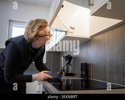 Ein männlicher Techniker untersucht den Kamin, während er einen Laptop in der Küche zu Hause hält Stockfoto