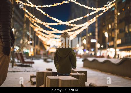 Rückansicht eines Kleinkindes in warmer Kleidung auf der beleuchteten Straße in der Stadt während der Nacht Stockfoto