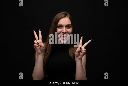 Junge Frau lächelt beim Friedenssieg V-Zeichen Geste mit beiden Händen vor schwarzem Hintergrund Stockfoto
