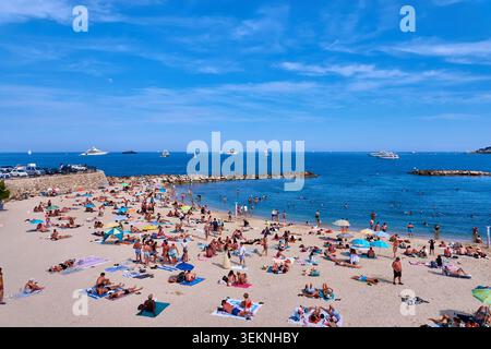 Überfüllter Sommerstrand in Antibes mit Sonnenanbetern, Sonnenschirmen und Booten an einer sonnigen Mittelmeerküste Stockfoto