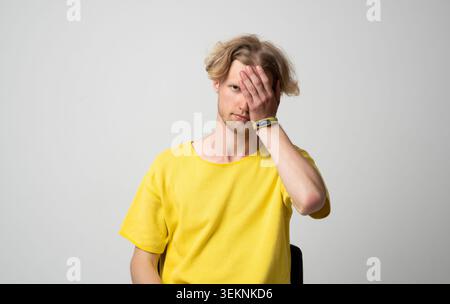 Junger Mann, der ein gelbes T-Shirt trägt, das teilweise sein Gesicht mit der Hand bedeckt und Konzepte von Scham, Verstecken, Privatsphäre, Emotionen, Geheimnis vermittelt. und Stockfoto