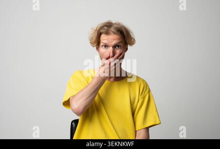 Junger Mann, der seinen Mund mit einer Hand bedeckt, starke Emotionen wie Schock, Überraschung, Unglauben vermittelt und ein Geheimnis bewahrt, mit breiten Augen Stockfoto