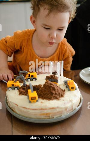 Ein glückliches Kind, ein dreijähriger Junge, bläst Kerzen auf der Geburtstagskuche aus. Das Baby sieht zufrieden und vollendet aus. Der Bastelkuchen ist auf der Geburtstagsparty der Familie zu sehen. Stockfoto