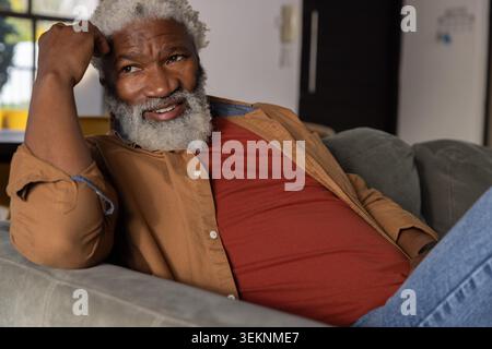 Reifer afroamerikanischer Mann, der sich auf dem Sofa in einem braunen Hemd über rostrotem T-Stück zurücklehnt und auf Pflanzgefäße blickt. Senior, Couch, Tageslicht, grau, gemütlich, Zuhause, Pflanze Stockfoto