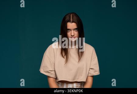 Eine junge Frau mit ernstem, intensivem Ausdruck, die ein beigefarbenes Oberteil trägt und vor einem minimalen blaugrünen Hintergrund steht Stockfoto