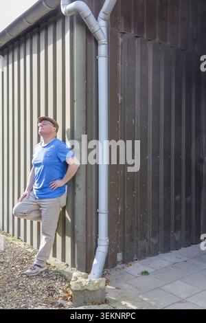 Erwachsener Mann, der sich in der Sonne sonnt, sich ruhig an die Blechwand lehnt, mit einem Fuß nach oben blickt, eine Pause einlegt oder eine Pause einlegt, um den Moment zu genießen Stockfoto