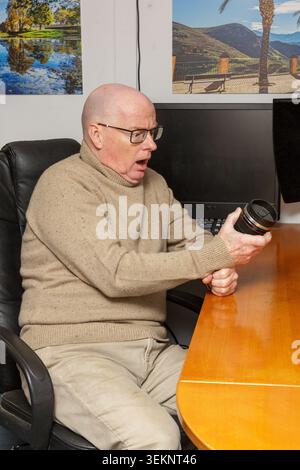 Ein Erwachsener Fotograf mittleren Alters mit einem überraschten Gesichtsausdruck und offenem Mund, hielt ein Objektiv und saß in einem Bürostuhl im Fotostudio dre Stockfoto