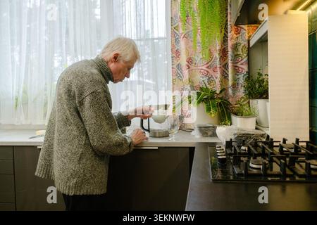 Ein Senior mit gebogenem Rücken gießt heißen Tee aus dem Wasserkocher in einen Becher. Das Bild erfasst die physischen Gegebenheiten des Alterns und den Komfort der Routine. Großvater in der Küche. Stockfoto