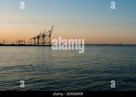 Hafenkrane und Pier Silhouetten bei Sonnenuntergang mit warmem Licht über dem Meer, die den industriellen Charme der Uferpromenade in Dämmerungsfarbe einfangen Stockfoto