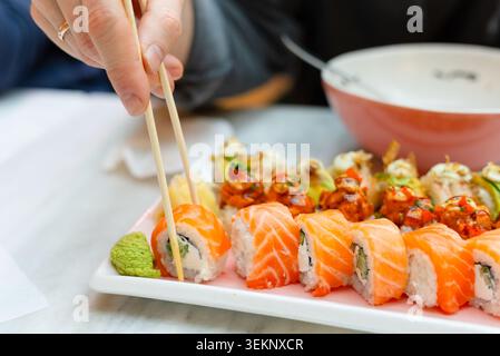 Eine Handperson hält Essstäbchen über einem Teller mit frischen Sushi-Brötchen in einem Restaurant. Diner wählt ein Stück Lachsnigiri aus der Sorte. Asiatische Küche mit Suppenschüssel im Hintergrund Stockfoto