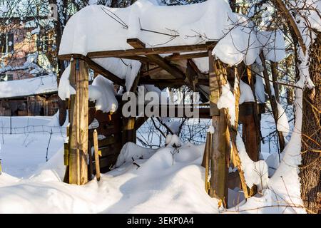Alte Holzkonstruktion, die nach einem schweren Wintersturm unter starkem Schnee vergraben wurde, und Sonnenlicht beleuchtet die dicken weißen Schichten, die den verfaulenden Gartenschacht bedecken Stockfoto