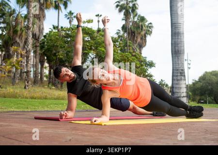 Eine fitte Frau in einem orangefarbenen Tanktop und ein Mann in schwarzer Sportkleidung üben seitliche Plank-Übungen auf Yogamatten, wobei der Schwerpunkt auf der Stabilität des Kerns während eines Ou liegt Stockfoto