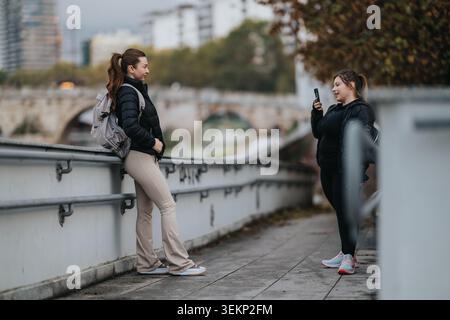 Zwei Frauen in Sportkleidung plaudern auf einer Brücke am Fluss, während eine ein Foto mit ihrem Handy macht Stockfoto