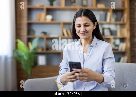 Junge Frau, die auf einem Sofa sitzt, lächelt, während sie ein Smartphone hält und mit sozialen Medien oder Nachrichten interagiert, die alltägliche digitale Kommunikation und Freizeit zu Hause repräsentieren Stockfoto