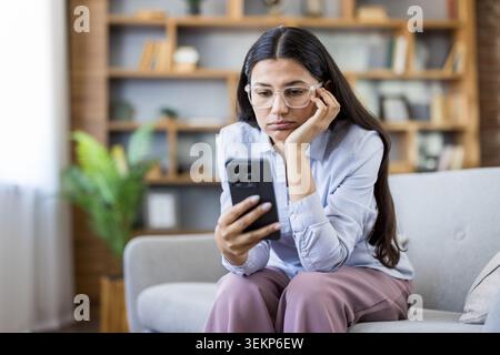 Junge Frau, die auf dem Sofa sitzt und ein Smartphone hält, traurig und enttäuscht aussieht, während sie Informationen liest, Gefühle von Einsamkeit und Angst erlebt Stockfoto