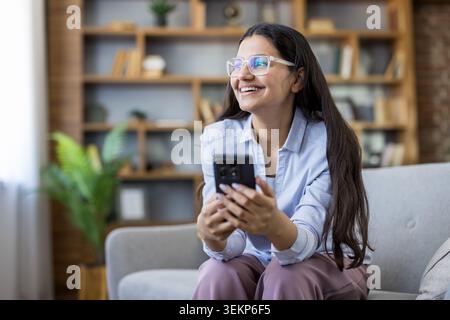 Eine junge Frau in Brille und blauem Hemd lächelt, während sie sich auf einem Sofa entspannt, ein Smartphone in einem modernen Home Office hält, Apps durchsucht und den Komfort ferngesteuert genießt Stockfoto