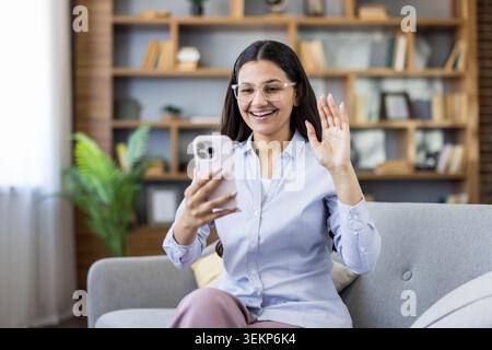 Junge Frau, die zu Hause auf einem Sofa sitzt, lächelt und winkt während eines Videoanrufs mit dem Smartphone, entspannt und fröhlich, während sie sich online mit Freunden oder Familie in einem gemütlichen Wohnzimmer verbindet Stockfoto
