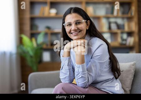 Eine junge indianerin, die eine klare Brille und ein Hemd trägt, in die Kamera blickt und lächelt, entspannt auf einer Couch sitzt und die Hände vor ihr in einem Zuhause oder Büro gefesselt haben Stockfoto