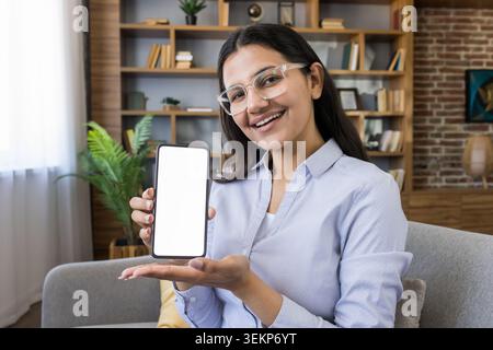 Eine junge Frau mit Brille lächelt in die Kamera, hält ein Smartphone mit leerem Bildschirm senkrecht in der einen Hand und präsentiert es mit der anderen Hand, was Raum für Werbung oder Inhalte bietet Stockfoto