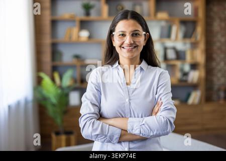 Die junge indische Geschäftsfrau in klarer Brille und hellblauem Hemd steht selbstbewusst mit den Armen, die in einem modernen Büro von einem Bücherregal gekreuzt werden, lächelnd und projiziert Professionalität und Erfolg Stockfoto