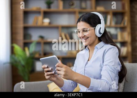 Junge Frau mit Brille und Kopfhörern, die sich auf dem Sofa entspannt und lächelt, während sie zu Hause Videos und Musik auf einem Tablet streamt, um Unterhaltung, Lernen oder E-Learning zu genießen Stockfoto