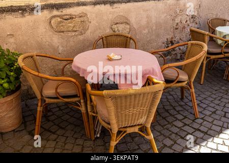 Eine Außenaufnahme einer bezaubernden europäischen Restaurantterrasse auf Kopfsteinpflaster, die bequeme Korbsessel um eine kleine Tischdecke zeigt Stockfoto