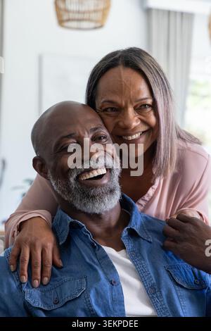 Seniorenpaar lacht und umarmt im Wohnzimmer, Mann trägt Denim-Hemd, Rattan-Anhänger Stockfoto