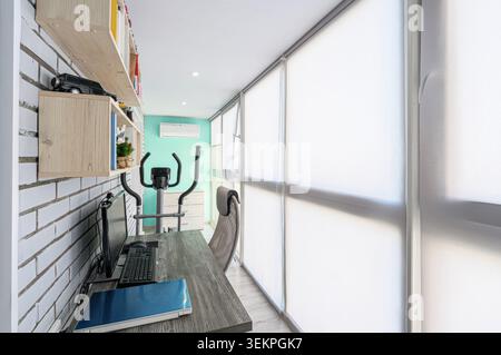 Modernes Heimbüro und Fitnessraum. Mit Schreibtisch, Computer, Bücherregal, Crosstrainer und großem Fenster mit Jalousien. Die Farbpalette ist hell und A Stockfoto