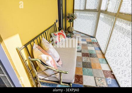 Gemütlicher Sonnenzimmer mit Bank, Kissen und einer Vase mit Blumen, farbenfrohen Fliesenböden und gemusterten Fensterschirmen Stockfoto