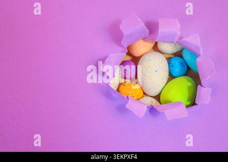 Wunderschöne bunte Ostereier, die durch lila zerrissenes Papier schauen, leuchtend farbige Ostereier-Leckereien auf einer lila flachen Lage, die Ostern feiern Stockfoto