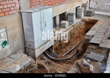 Erdkabel, die während der Straßenbauarbeiten in einem tiefen Graben unter einem Elektroverteilerkasten freiliegen. Stockfoto