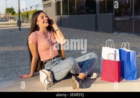 Junge Frau in lässiger Kleidung, die an einem Sommernachmittag lächelnd sitzt und mit mehreren Einkaufstaschen telefoniert. Sommerverkauf und Lifestyle. Stockfoto